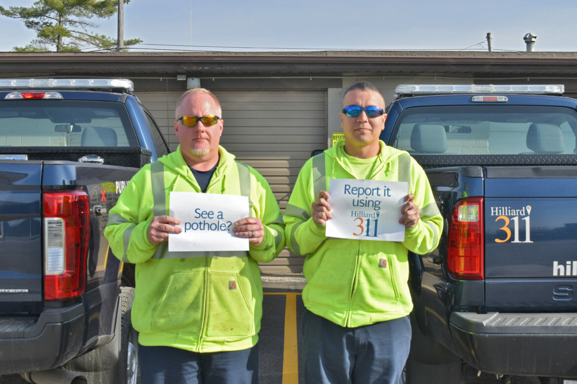 Public service workers holding up signs saying "See a pothole? Report it using Hilliard 311".