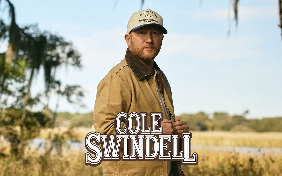 A man in a jacket and baseball hat stands at the edge of a field