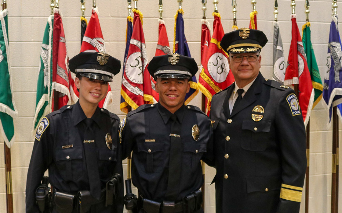 New Hilliard Police Officers Alexa Napier and Carter Ely stand beside Chief of Police Michael Woods in front of class flags at the Columbus Police Training Academy.